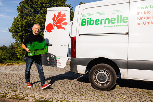 KI generiert: Ein Mann trägt eine grüne Kiste von einem Lieferwagen mit der Aufschrift "Bibernelle".