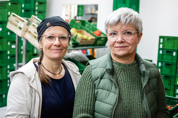 KI generiert: Zwei Frauen stehen in einem Raum mit Kisten und Obst im Hintergrund.